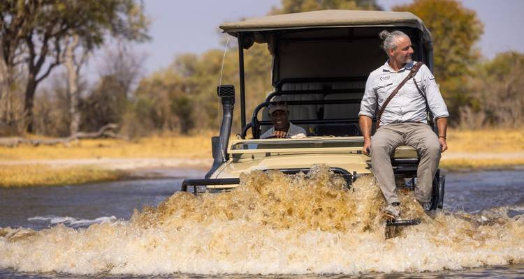Mann, der auf dem Rand eines Safari-Fahrzeugs durch Wasser fährt.