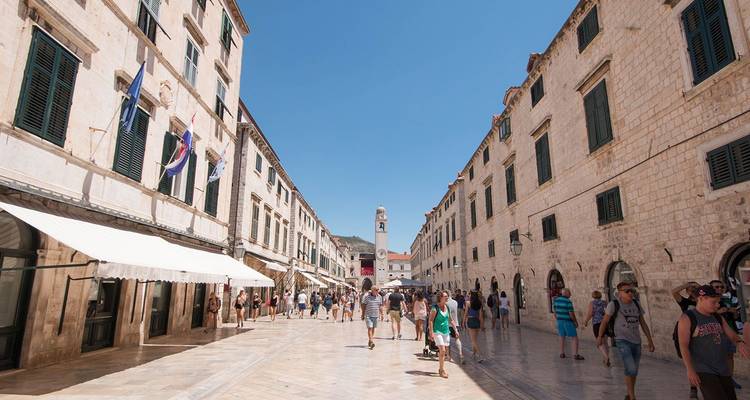 Voetgangers die op een zonnige zomerdag over de marmeren Stradun-straat van Dubrovnik flaneren, omgeven door stenen gebouwen.