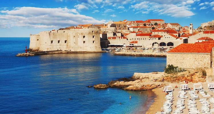 Uitzicht over de rustige baai naar het fort en de oude stad van Dubrovnik met een klein strand en ligbedden op de voorgrond.