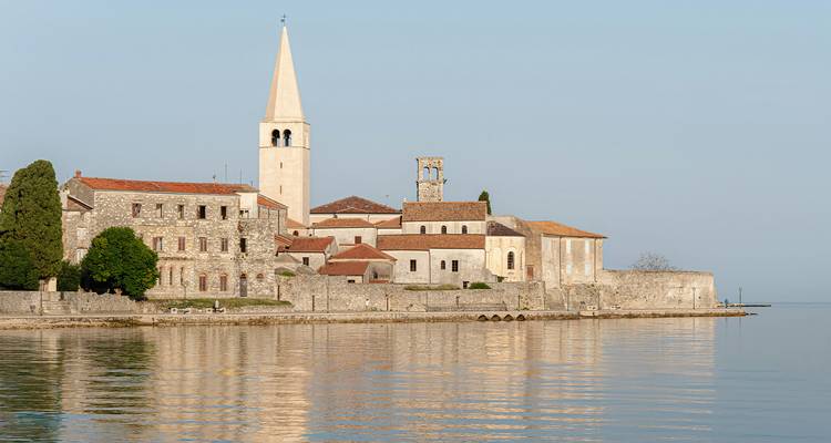 Stenen basiliek en slanke klokkentoren staan aan een kalme Adriatische kustlijn bij zonsondergang in Poreč.