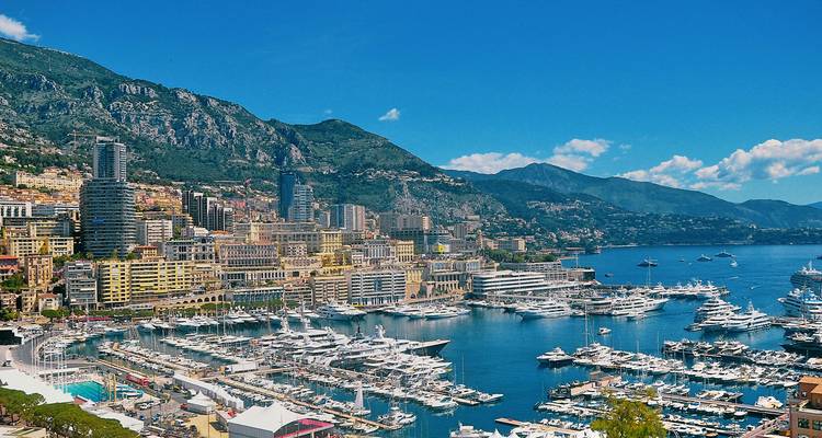Panorama ensoleillé du paysage urbain en terrasses de Monaco et du port rempli de yachts encadrés par les montagnes.