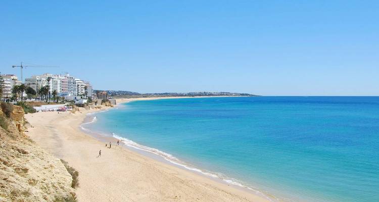 Largo tramo de playa dorada del Algarve que se encuentra con un mar turquesa tranquilo y un pueblo resort a lo lejos