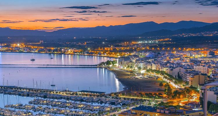 Ciudad costera de Roses iluminada al atardecer con puerto deportivo y montañas a lo lejos