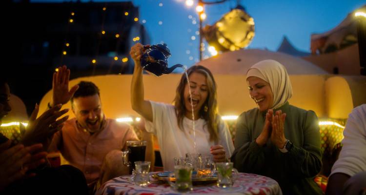 Los amigos comparten risas mientras una mujer vierte teatralmente té desde lo alto sobre pequeños vasos en la cubierta de un barco al anochecer en el Nilo.