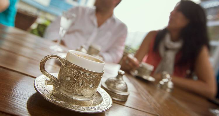 Nahaufnahme einer kunstvollen türkischen Kaffeetasse auf einem Holztisch mit unscharfen Gästen, die sich dahinter unterhalten.