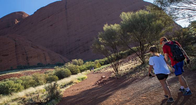 Pareja caminando por un sendero del desierto hacia las enormes cúpulas de Kata Tjuta en el Parque Nacional Uluru-Kata Tjuta