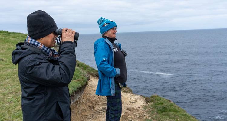 Dos individuos disfrutando de una vista costera mientras uno usa binoculares.