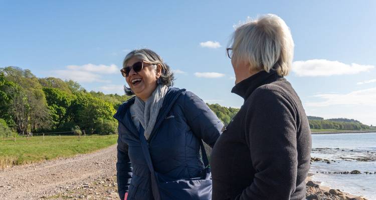 Dos mujeres disfrutando de una risa juntas junto al mar bajo un cielo azul despejado.