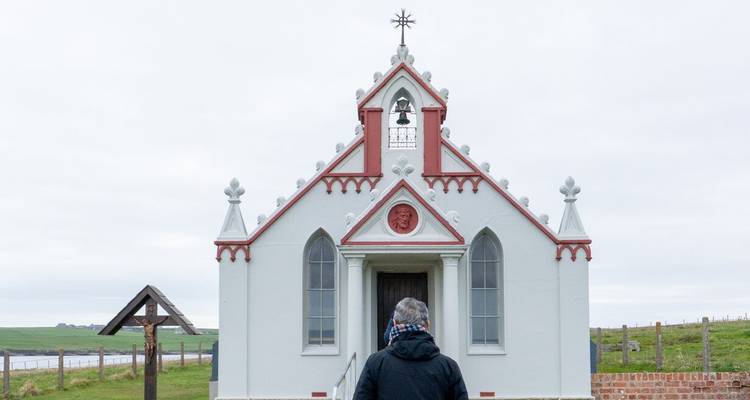 Una persona acercándose a una pequeña capilla blanca con acentos rojos bajo un cielo nublado.