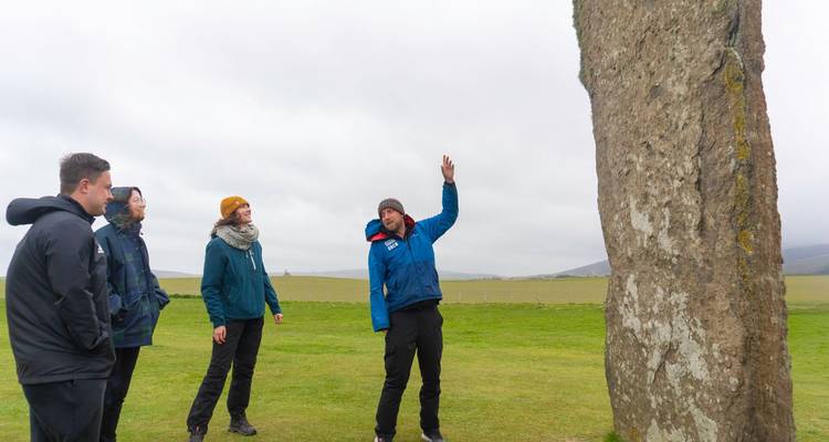 Ein Reiseleiter deutet auf einen aufragenden Menhir, während eine kleine Gruppe an einem bewölkten Tag zuhört.