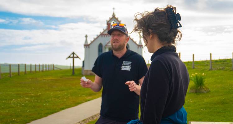 Zwei Reisende unterhalten sich außerhalb einer kleinen weiß-roten Kapelle, die sich in einer hügeligen Landschaft befindet.