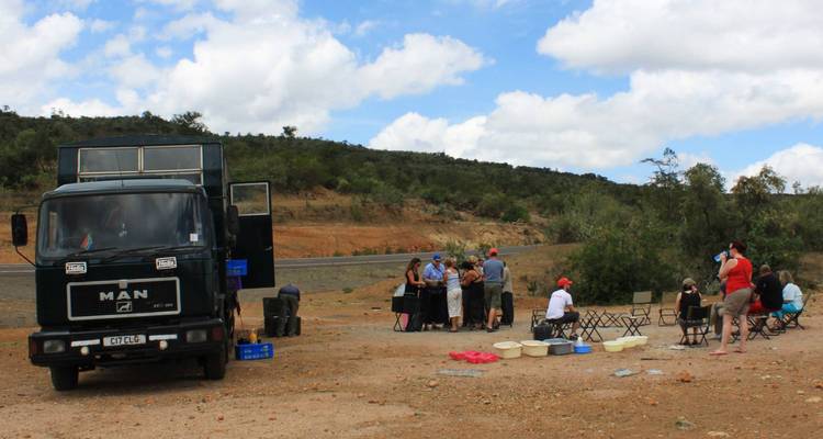 Camión de expedición terrestre estacionado al lado del camino mientras un grupo de viajeros prepara el almuerzo en mesas y sillas plegables.