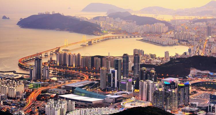 Stadtlandschaft mit Brücke über Wasser bei Sonnenuntergang, bergige Kulisse.