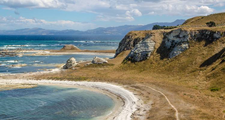 Schilderachtige kustrotsen en een gebogen zandstrand met turquoise water en verre bergen onder een heldere blauwe lucht.