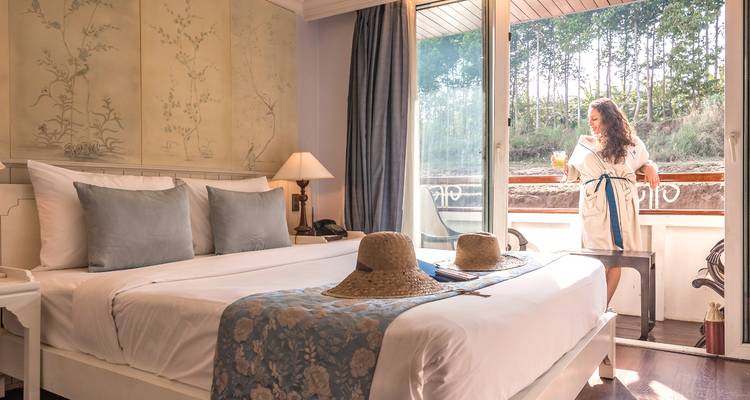 Hotelzimmer mit einer Person, die mit einem Getränk auf dem Balkon steht und auf einen Garten blickt.