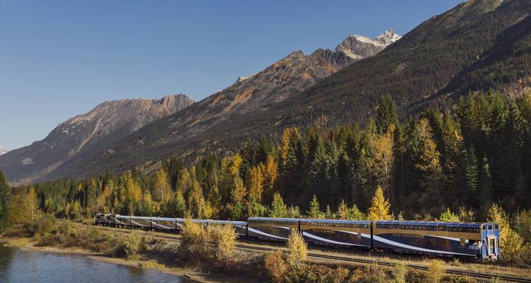 The long train winds beside a river through dense evergreen forest and jagged peaks.
