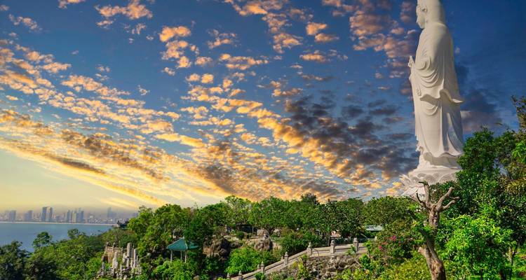 Standbeeld op een berg met een stad en oceaan op de achtergrond bij zonsondergang.