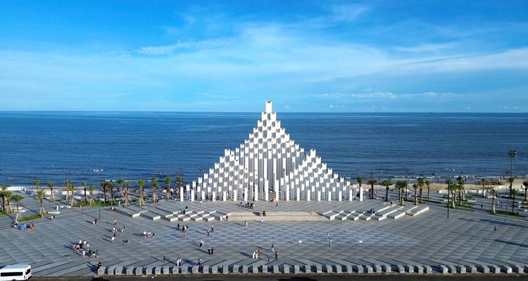Groot modern plein met een opvallend wit pyramidevormig monument naast een met palmen omzoomde kustlijn onder een helderblauwe hemel.