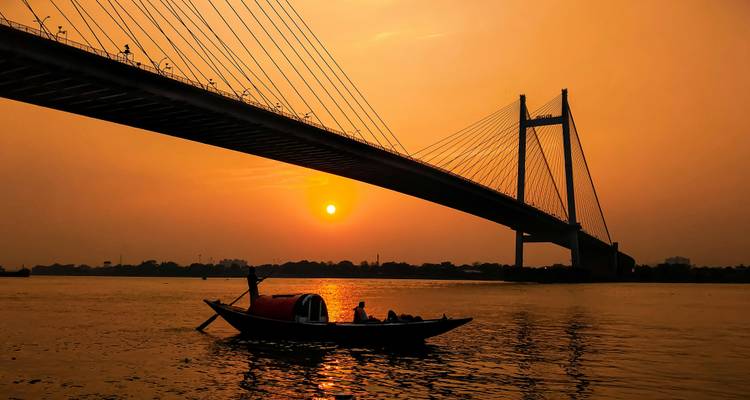 Un bateau en bois se dessine en silhouette glissant sous le Pont Vidyasagar à haubans dans un coucher de soleil orange flamboyant sur le fleuve Hooghly.