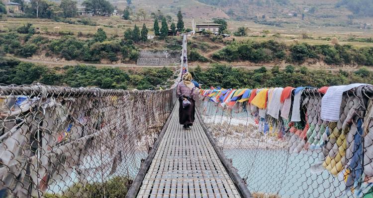 Une femme traverse un long pont suspendu bordé de drapeaux de prière flottants au-dessus d'une rivière turquoise.