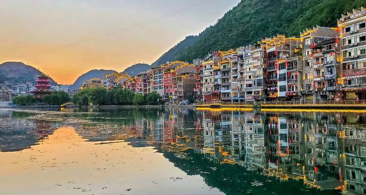 Pintoresco pueblo ribereño en Guizhou con casas tradicionales escalonadas iluminadas al atardecer reflejándose en aguas tranquilas.