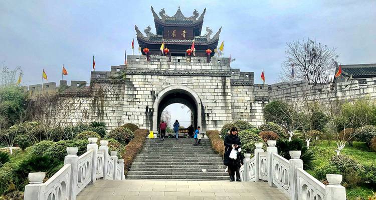 Puerta arqueada de piedra de una antigua muralla china a la que se accede por un puente blanco, con algunos visitantes caminando y una mujer posando enfrente en un día nublado.