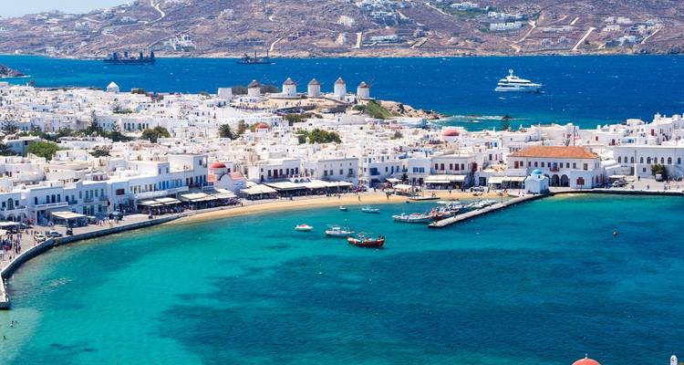 Aerial view of Mykonos town harbour, turquoise bay, and iconic windmills on the horizon.