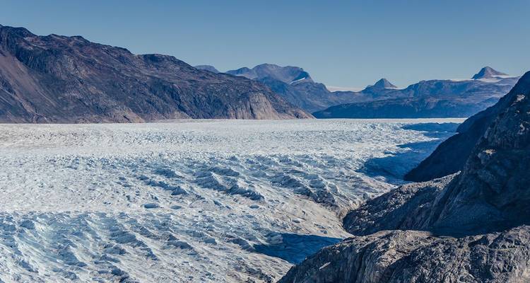 Immense langue glaciaire crevassée s'étendant entre des montagnes grises escarpées sous un ciel bleu dégagé.