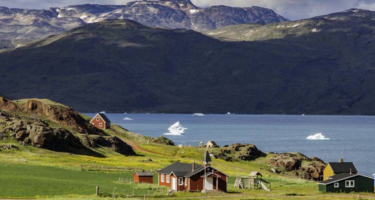 Établissement groenlandais tranquille avec des chalets rouges et une église situés dans des champs verdoyants à côté d'un fjord parsemé d'icebergs.
