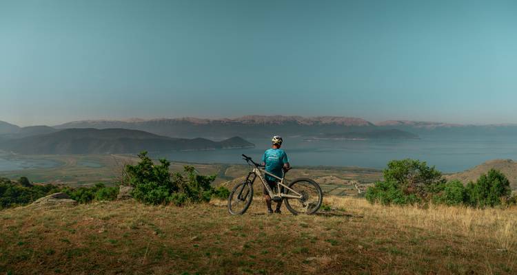 Cycliste de montagne debout avec son vélo surplombant un lac et une chaîne de montagnes lointaine.