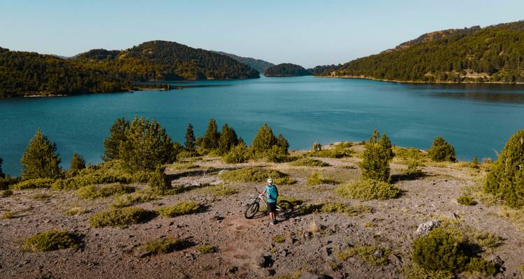 Vue aérienne d'un cycliste à côté d'un lac de montagne turquoise entouré de collines boisées.