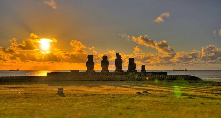 Estatuas moai recortadas contra una puesta de sol naranja vibrante sobre el Océano Pacífico.