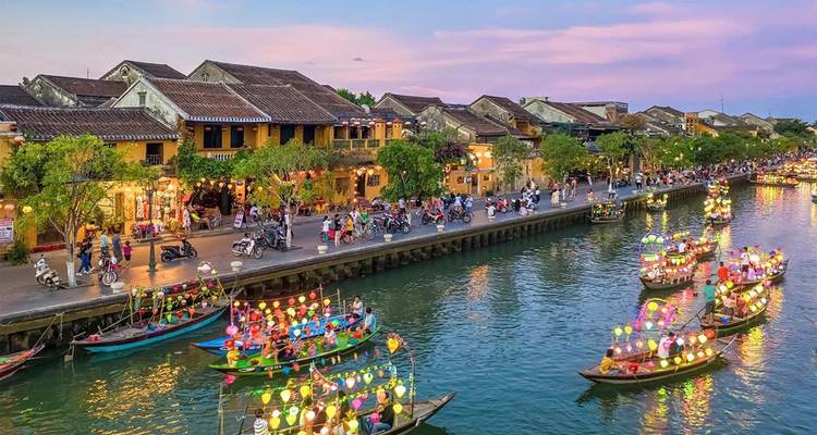 Scène de rivière à Hoi An avec des bateaux et des bâtiments illuminés par des lanternes au crépuscule.