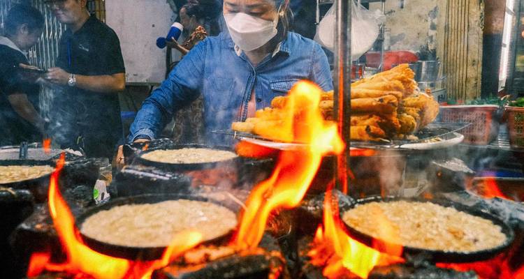 Food being cooked on open flames in a market setting, people wearing masks.