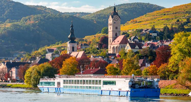 Een blauw-wit riviercruiseschip vaart langs terrasvormige herfstwijnagarden en een kerktoren in Oostenrijks Wachau-dal.