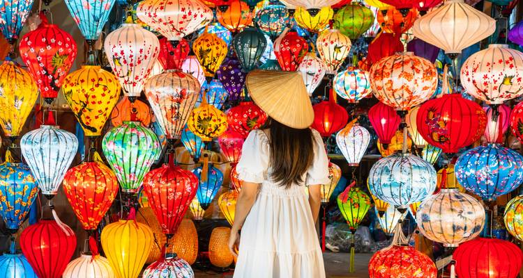 Eine Frau in einem weißen Kleid und mit konischem Hut steht zwischen bunten Laternen.
