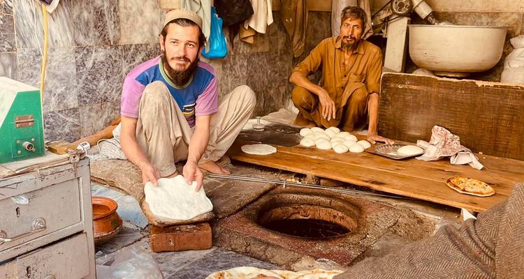 Dos panaderos preparando panes planos junto a un horno tandoor tradicional dentro de una tienda modesta