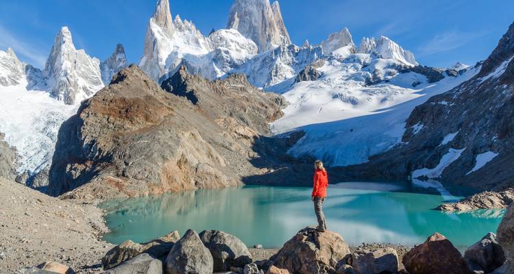 Eine Person in einer roten Jacke steht da und blickt auf einen Bergsee mit schneebedeckten Gipfeln.