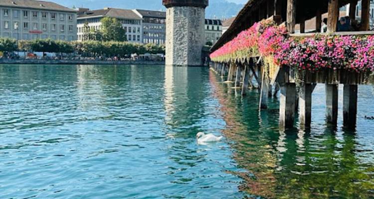 Puente histórico de madera cubierto con flores sobre un río con montañas al fondo.