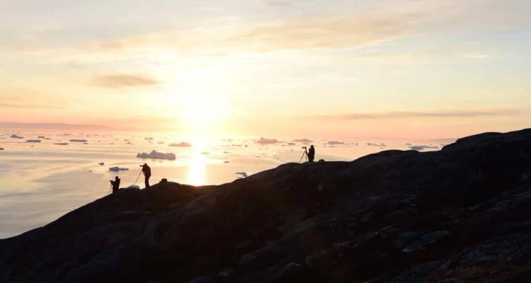 Silhouetten von Wanderern auf einem felsigen Grat mit Blick auf einen eisbergbedeckten arktischen Sonnenuntergang