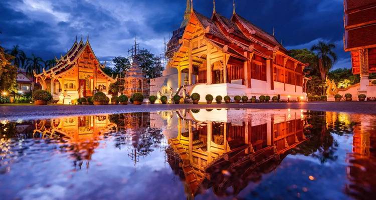 Illuminated Thai temples reflected in a rain puddle under dramatic dusk sky in Chiang Mai