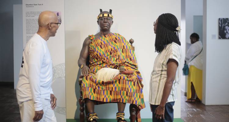 Des visiteurs conversent à côté d'un mannequin grandeur nature d'un roi Ashanti assis en robes royales kente dans une galerie de musée.