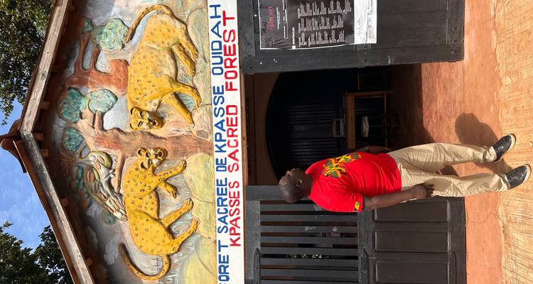 Un voyageur se tient devant le panneau d'entrée sculpté de la Forêt Sacrée d'Ouidah orné de reliefs de léopards peints.