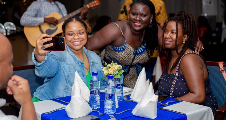 Freunde machen ein Selfie um einen Esstisch, der mit blauen Tischwäschen, Wasserflaschen und gefalteten Servietten gedeckt ist, während im Hintergrund ein Gitarrist spielt.