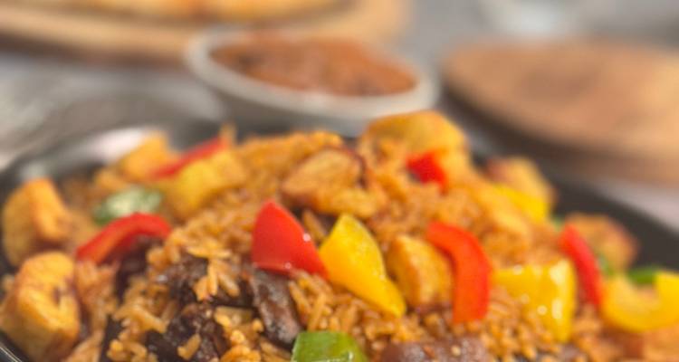 Nahaufnahme eines bunten Tellers mit Jollof-Reis, Paprika und Kochbananen in weicher Unschärfe.