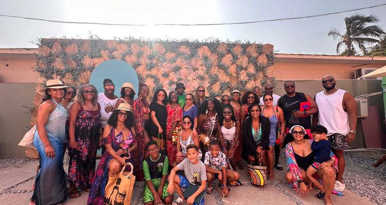 Große multikulturelle Reisegruppe posiert in Badeanzügen und Strandhüten vor einer dekorativen tropischen Kulisse.