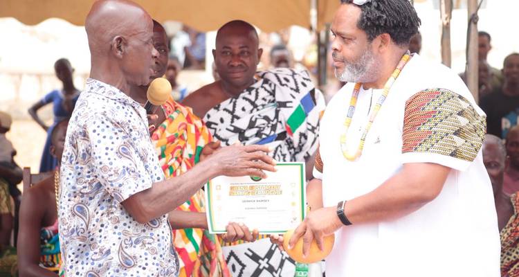 Gemeinschaftszeremonie, bei der ein Häuptling einem Besucher eine gerahmte Urkunde in traditioneller Kente-Umgebung überreicht.