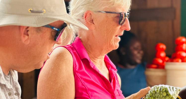 Reisende betrachten frische Produkte an einem lebendigen ghanaischen Marktstand mit roten Tomaten, die dahinter gestapelt sind.