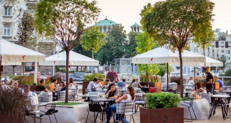 Personas cenando al aire libre en un café con árboles y edificios.