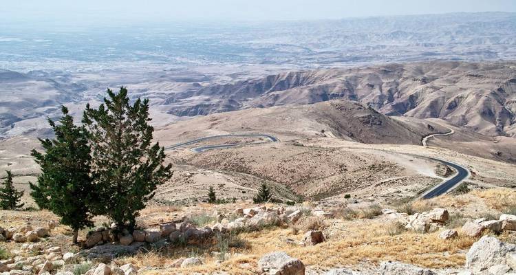 Une vue panoramique des terres hautes avec une route en asphalte sinueuse serpentant à travers des collines arides et accidentées vers une vallée lointaine.